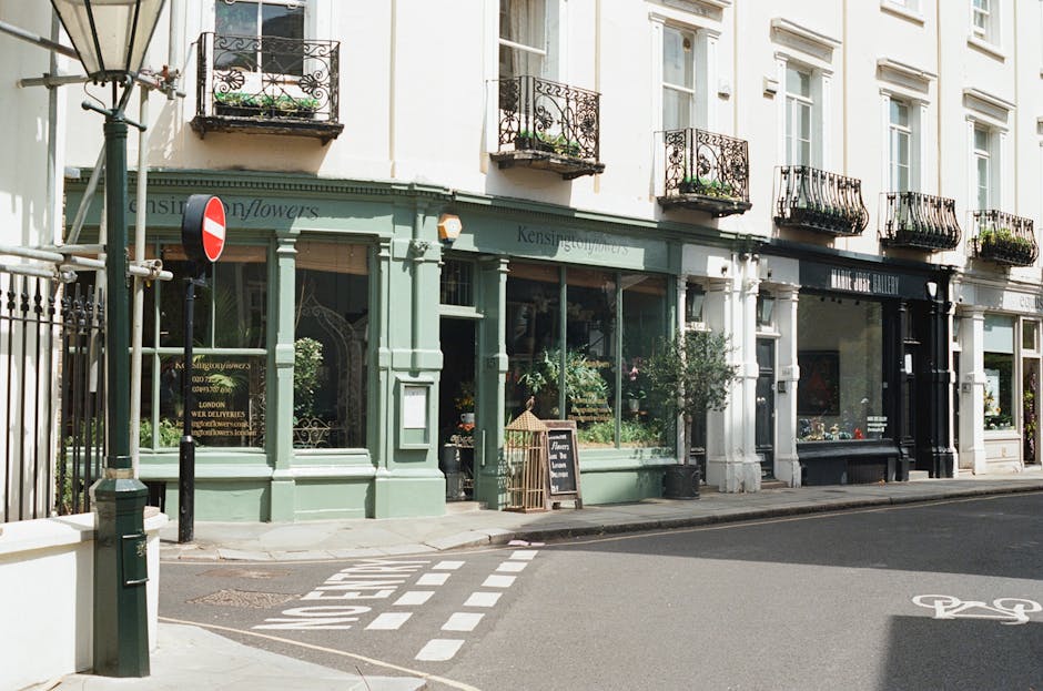 A street view featuring a row of charming, historic storefronts with large glass windows displaying an assortment of potted plants, floral arrangements, and garden ornaments. The building facades are