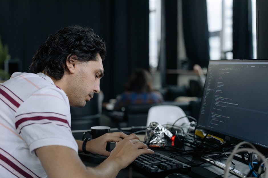 A young man with dark, wavy hair, wearing a white t-shirt with red and purple stripes, is seated at a desk working on a computer. His focus is on the monitor displaying lines of code or programming sc